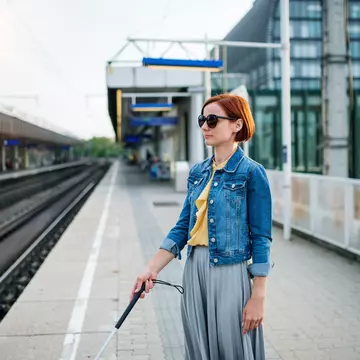 portrait of a blind woman with a white cane standing outside, waiting for public transport in summer.