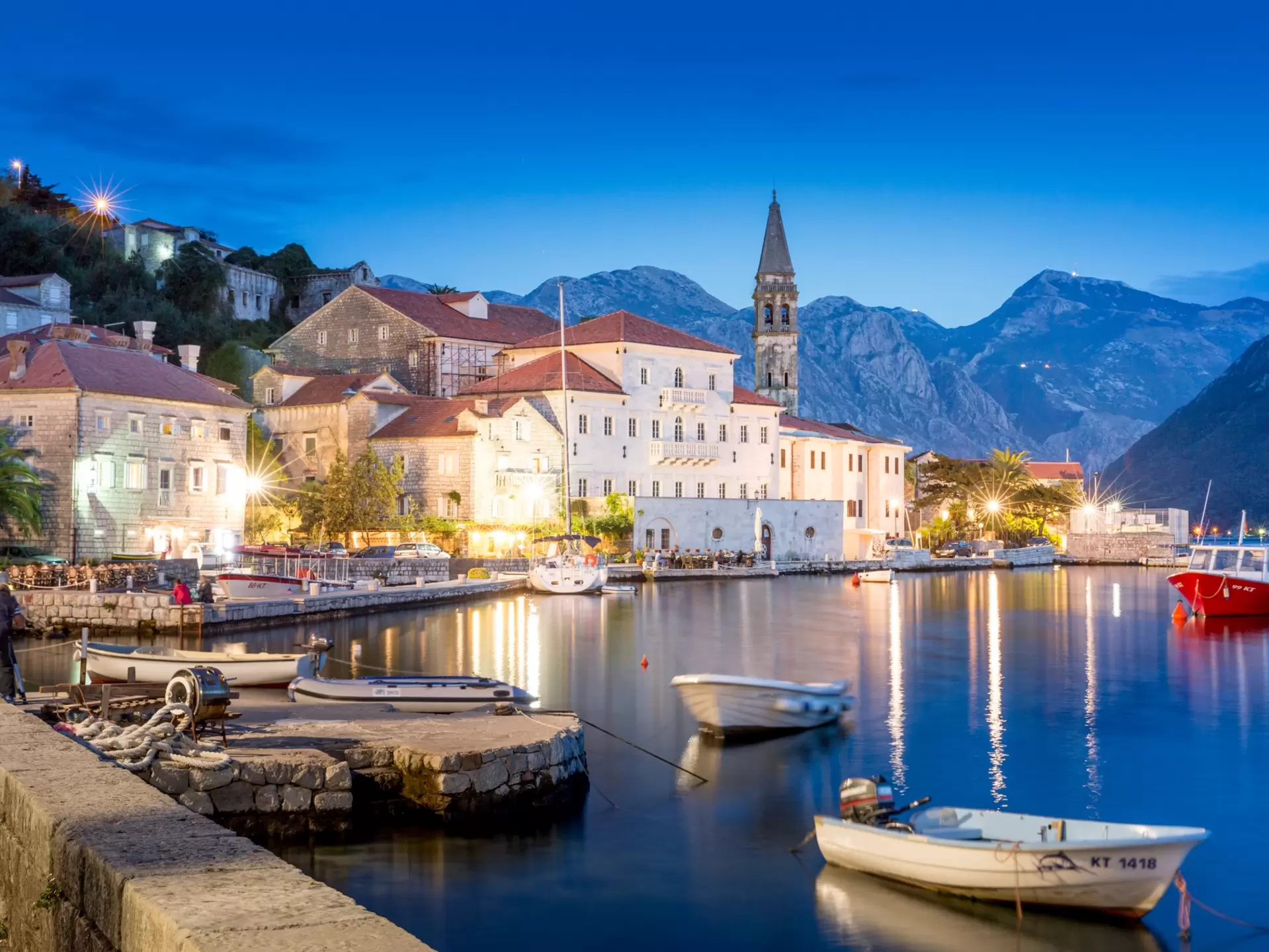 waterfront of a European town at night, with a church, small boats docked and mountains in the background