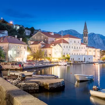 waterfront of a European town at night, with a church, small boats docked and mountains in the background
