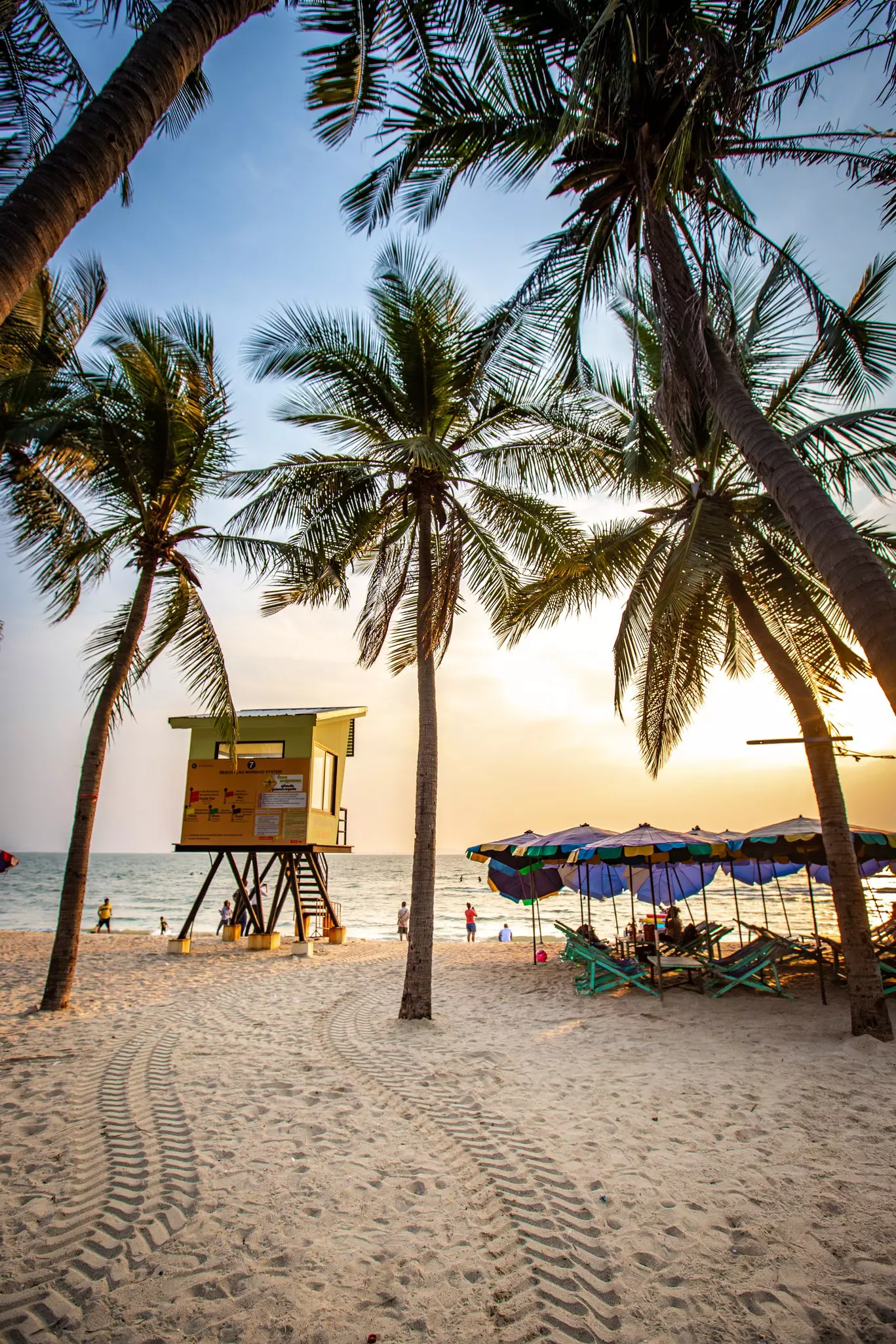 Tire tracks weave in the sand between two palm trees toward a lifeguard tower on a beach with umbrellas, chairs and another palm tree to the right of the tracks. People are on the shore in the distance.