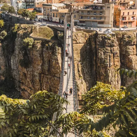 People walk on a narrow bridge across a deep gorge to a town on the other side.