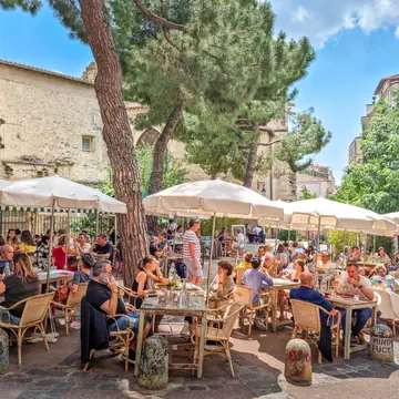 People sit at outdoors tables shaded by trees in a city square