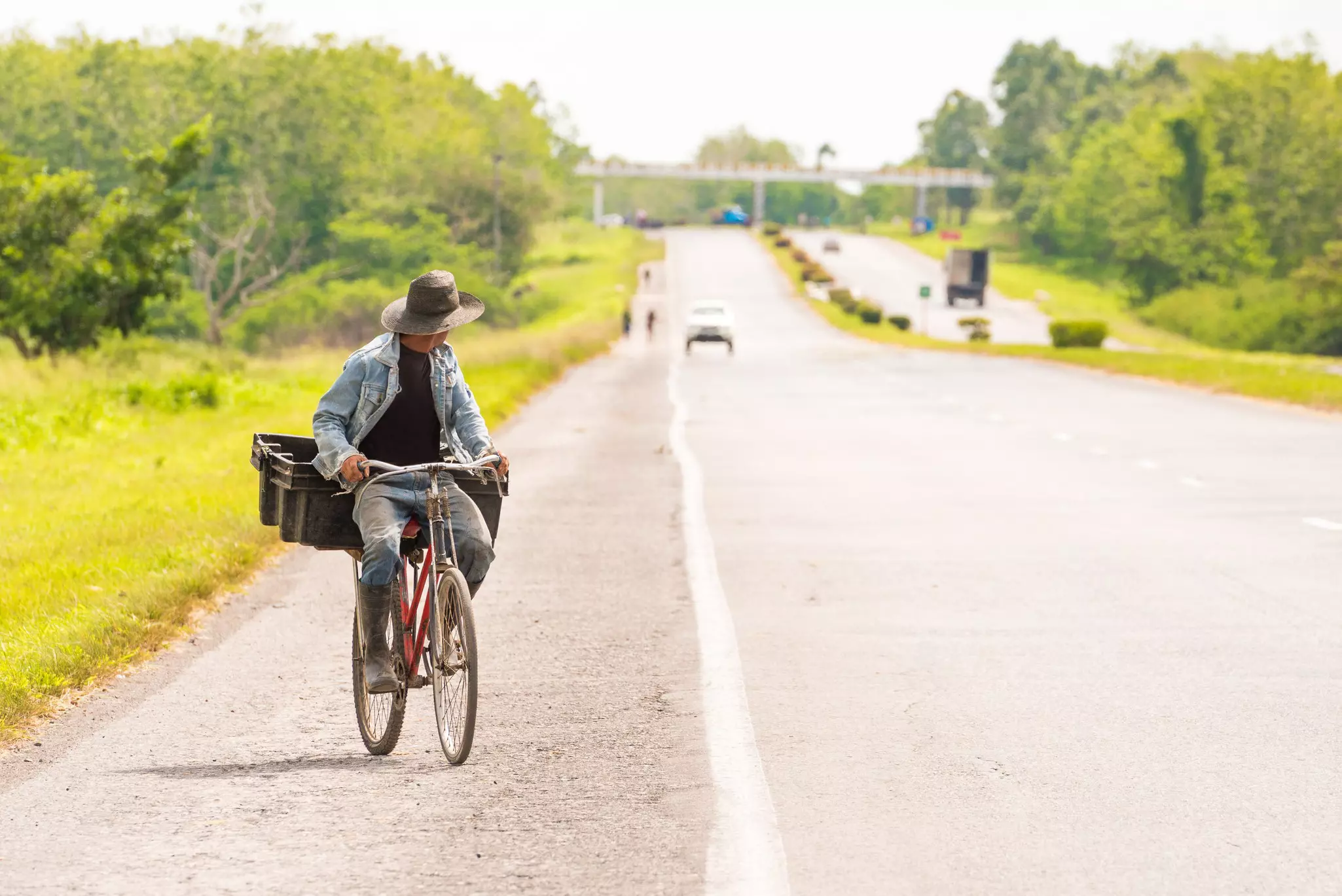 A man is riding along the road on a bicycle with a hat, looking behind him
