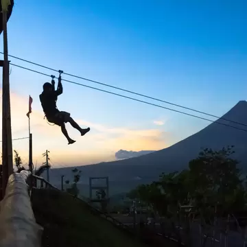 Silhouetted person on a zipline against a dusk sky, with a large symmetrical volcano rising beyond trees.