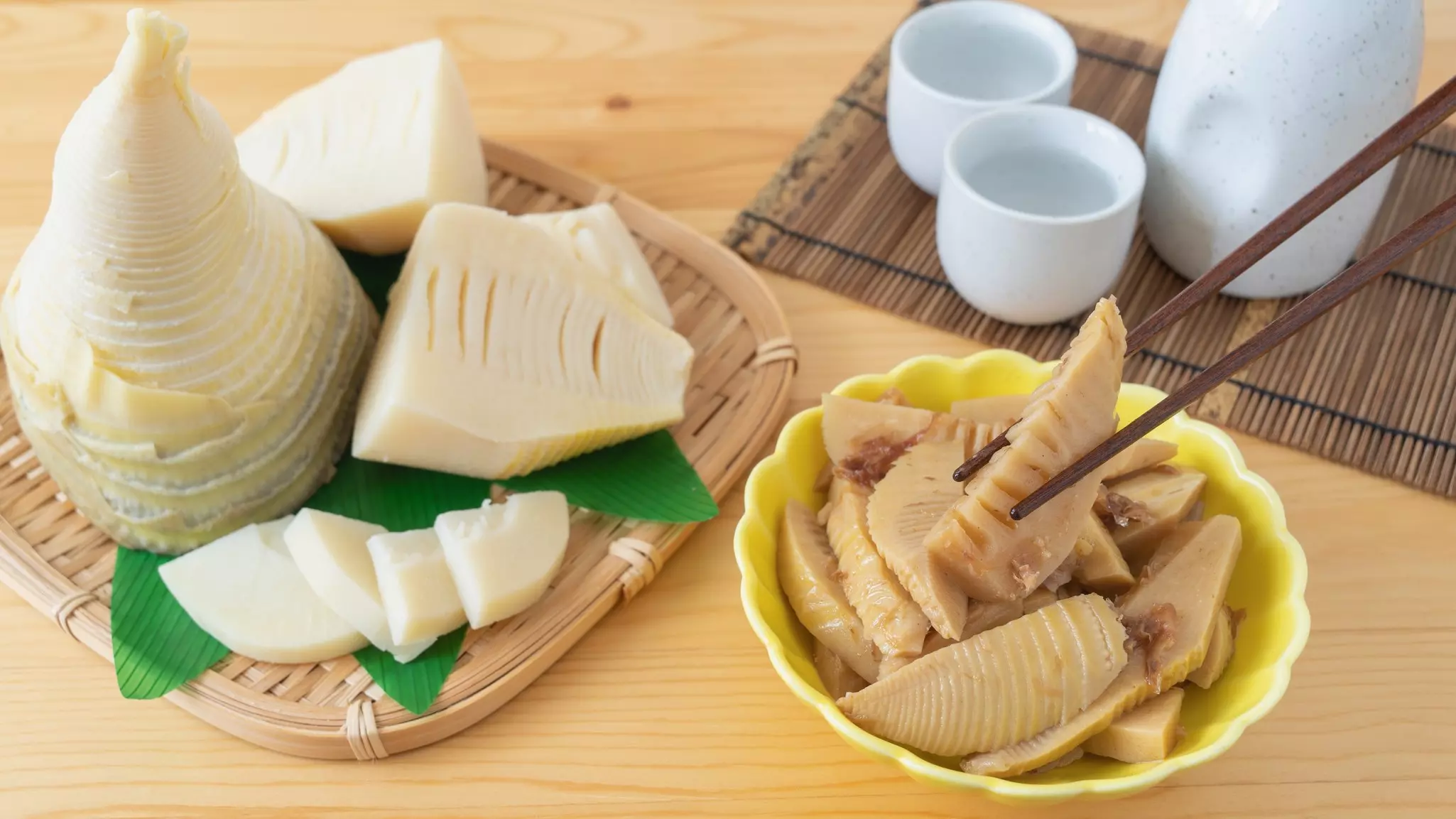 A small yellow bowl filled with sweet and spicy bamboo shoots. On the left is a raw bamboo shoot. In the background on the right is a white jug and two white cups filled with water.