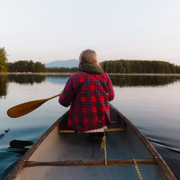 A person wearing a red plaid flannel shirt rows a canoe on a lake, with mountains in the distance.
