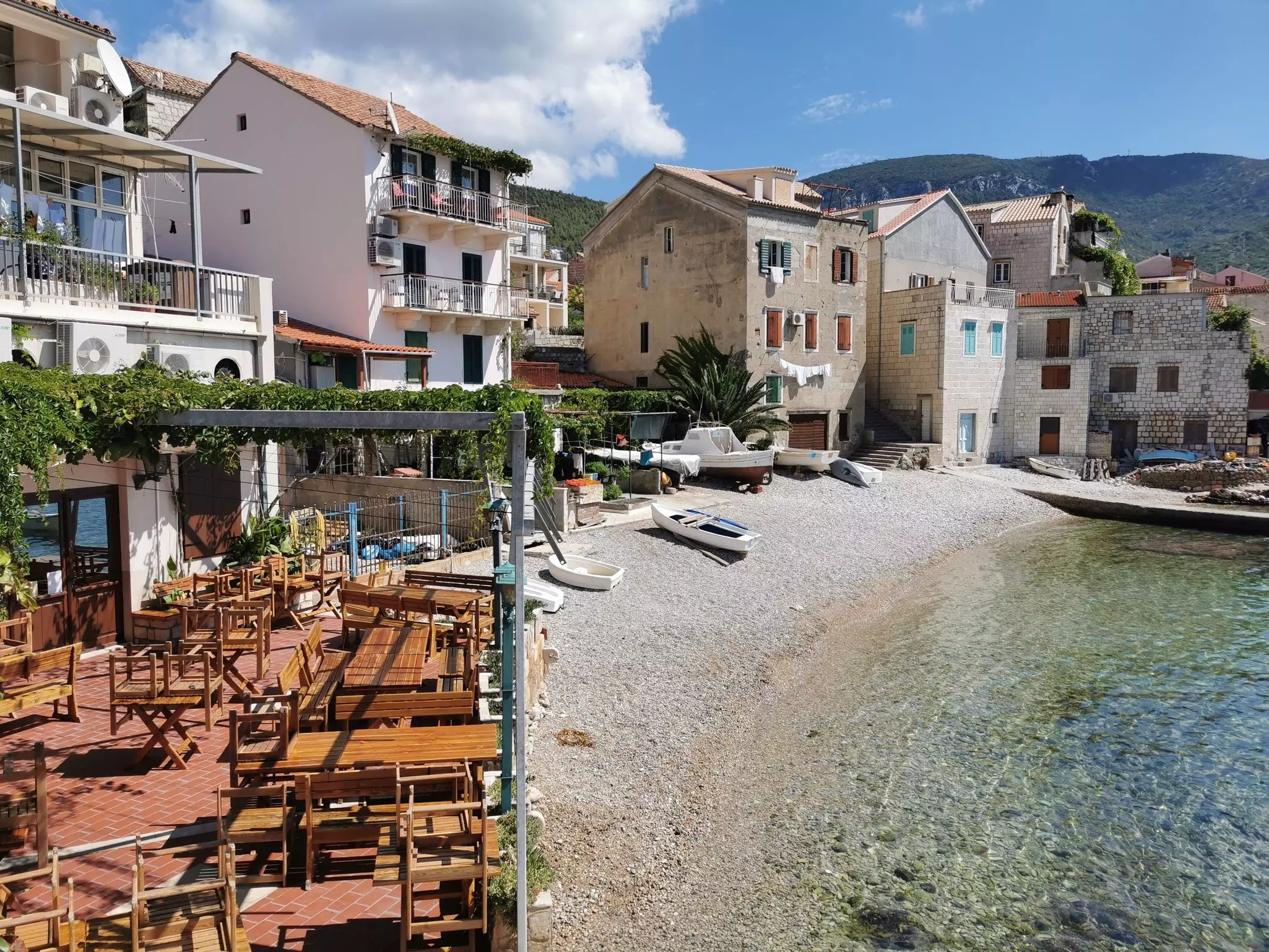 A pebble beach with boats pulled up onto it lined with a small restaurant with wooden tables and chairs and stone buildings in the background