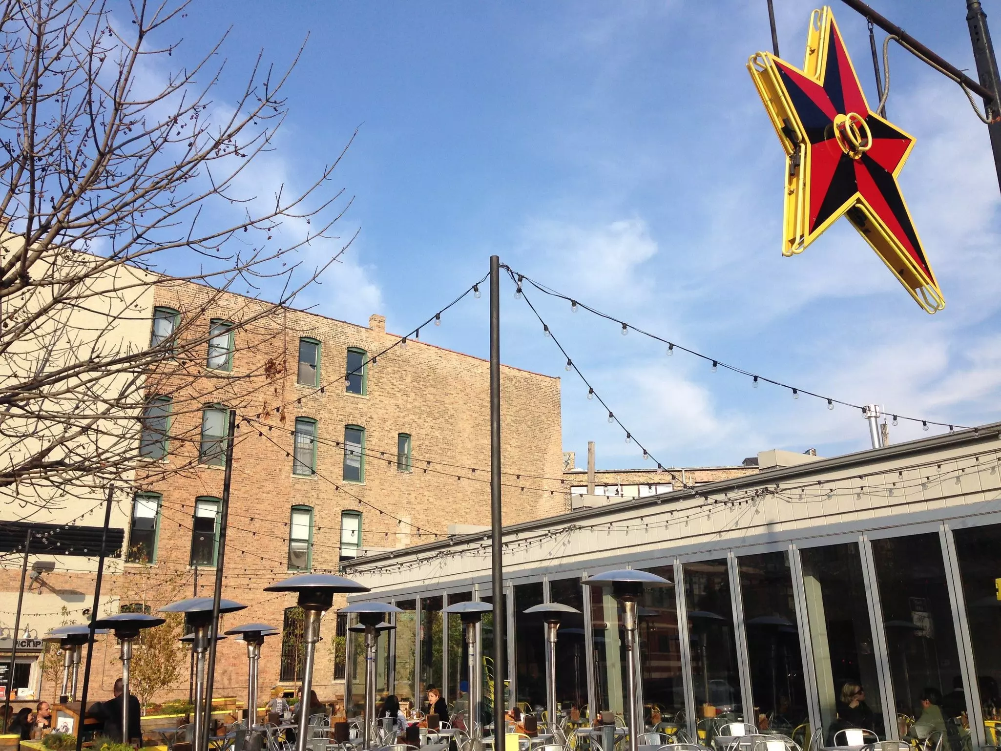 People eating outside a restaurant that has lights stringed over and heat lamps scattered around the dining area. A large black, red and yellow star hangs in the upper left corner.