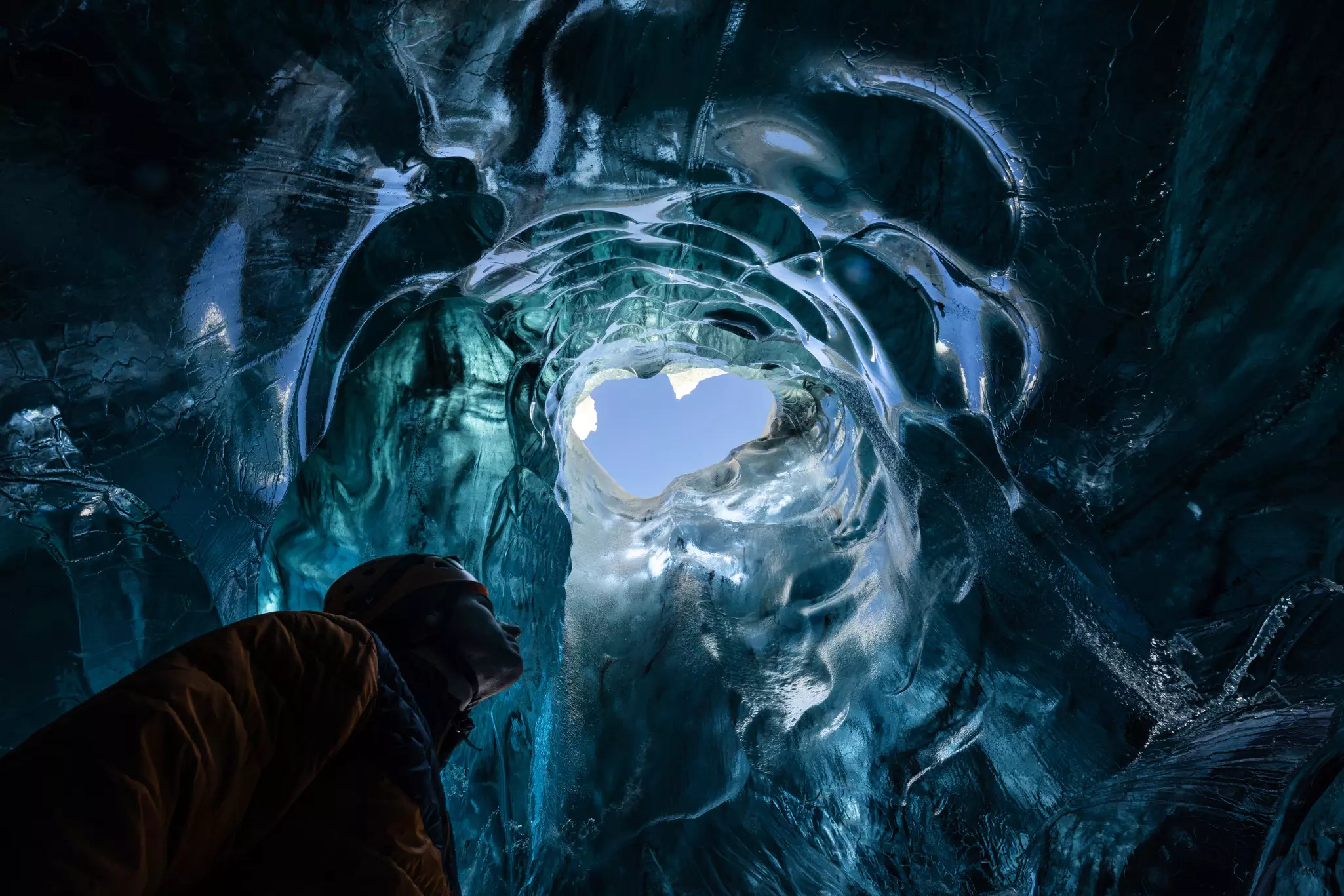 A view inside a glacier, looking up at the sky surrounded by walls of ice glimmering blue