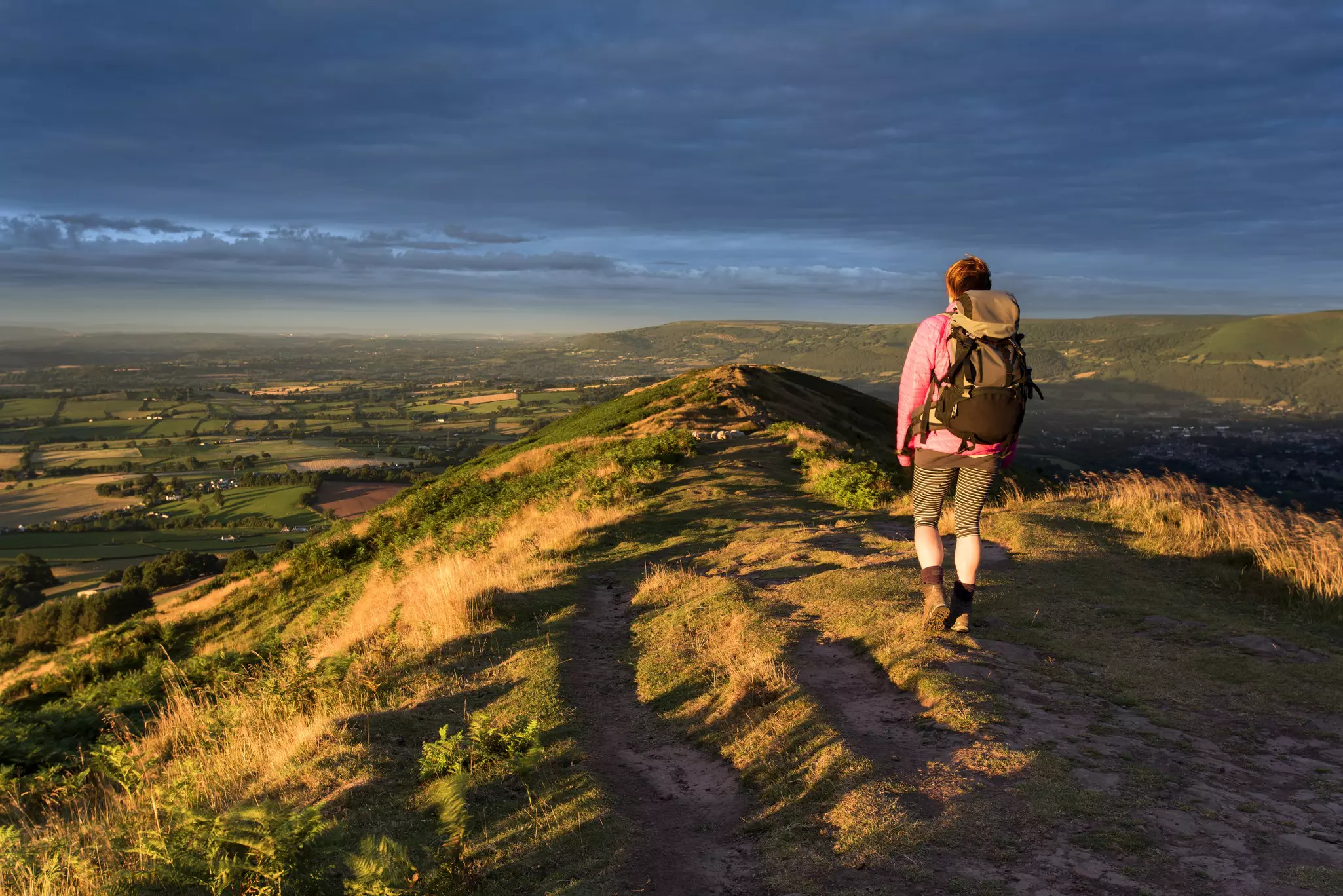 Woman hiker at sunrise with striped leggings, pink jacket and black-and-gray backpack walking along the ridge of a mountain with green fields and hills in the distance.