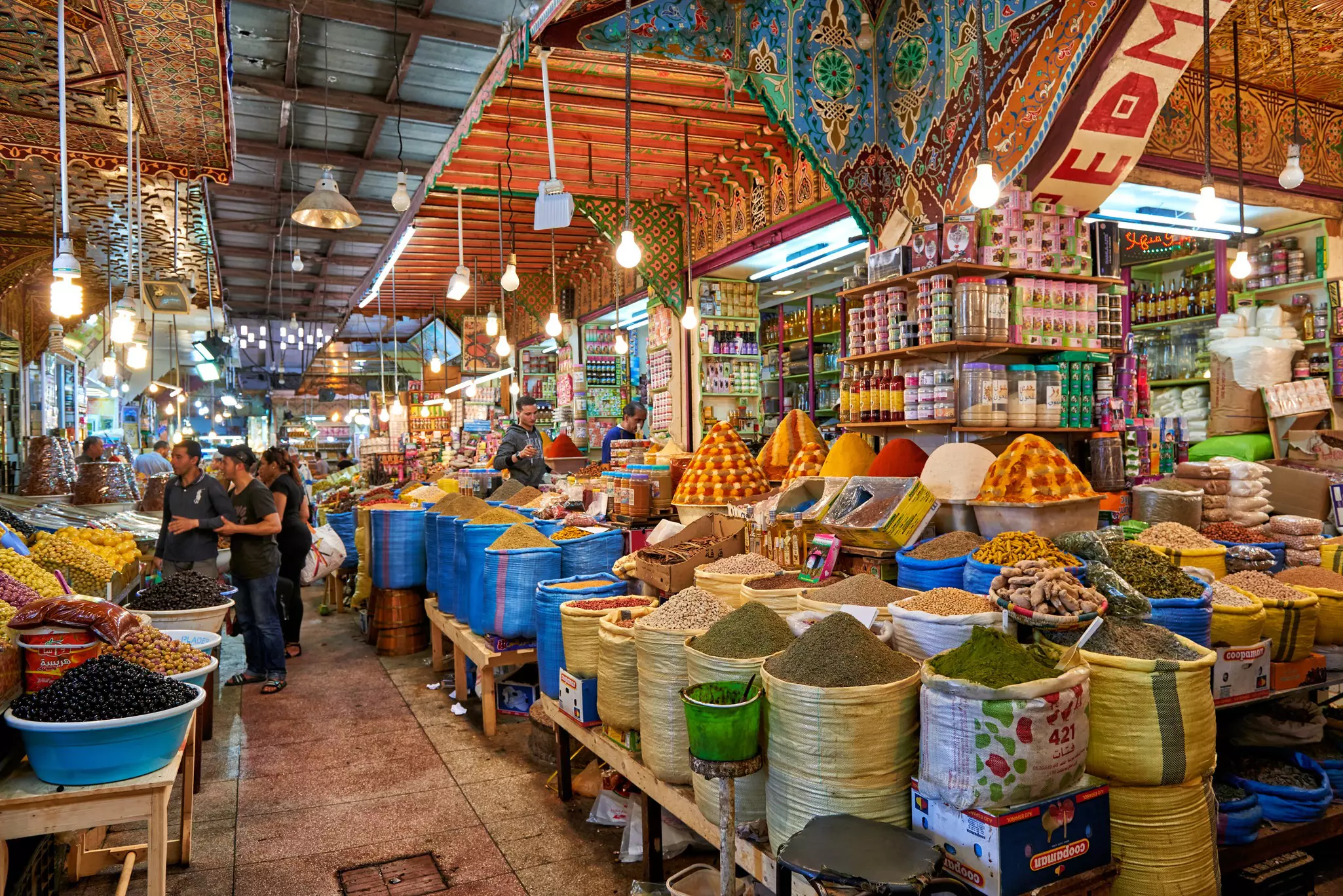 A covered market place with stalls selling spices and olives piled high in bags and baskets.