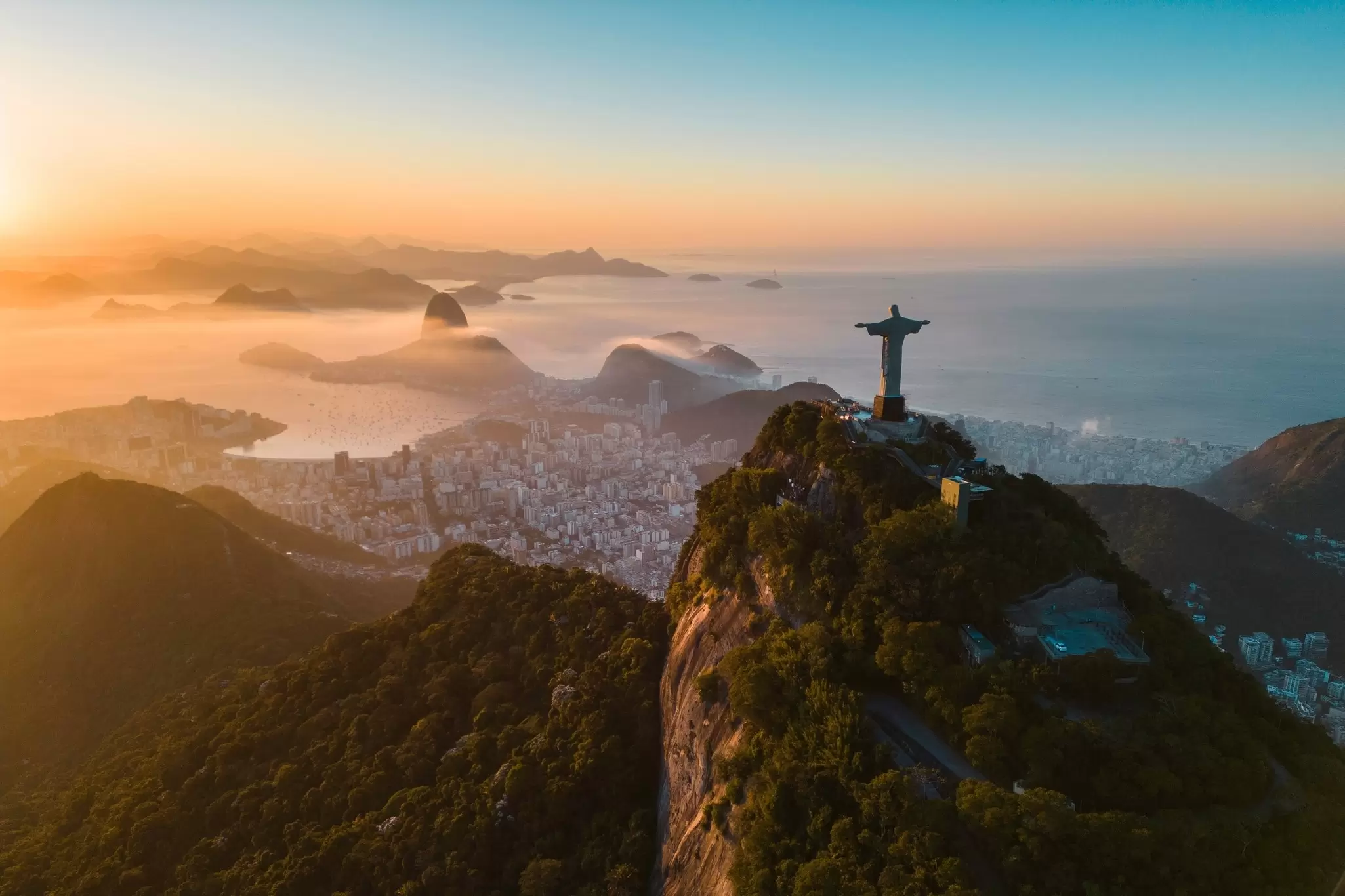 Sunrise on a statue with outstretched arms is on top of a mountain overlooking a city by the sea.