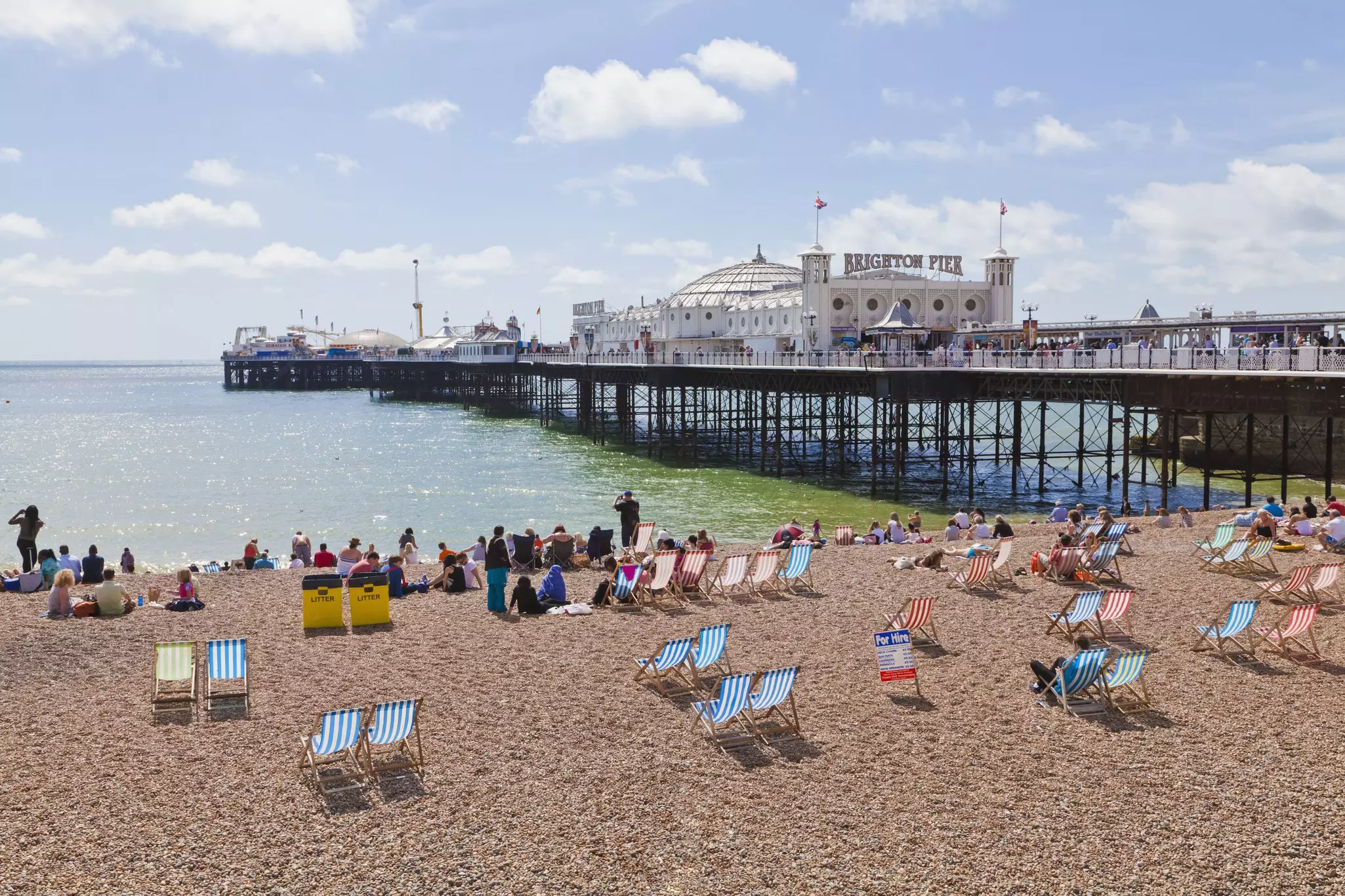 People sit on deckchairs on a pebbly beach with a large Victorian amusements pier stretching out into the ocean 