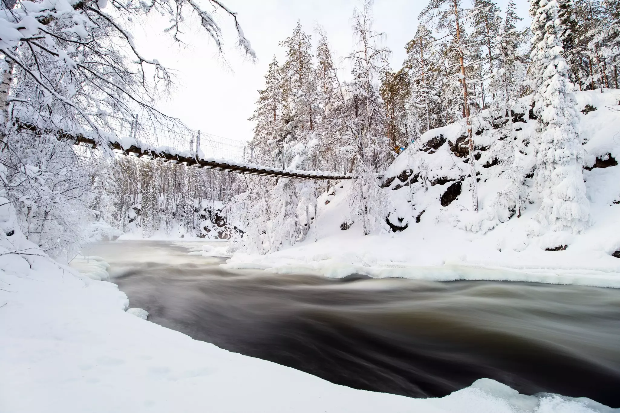 A rope bridge across a semifrozen river in a snowy forest landscape in Finland.