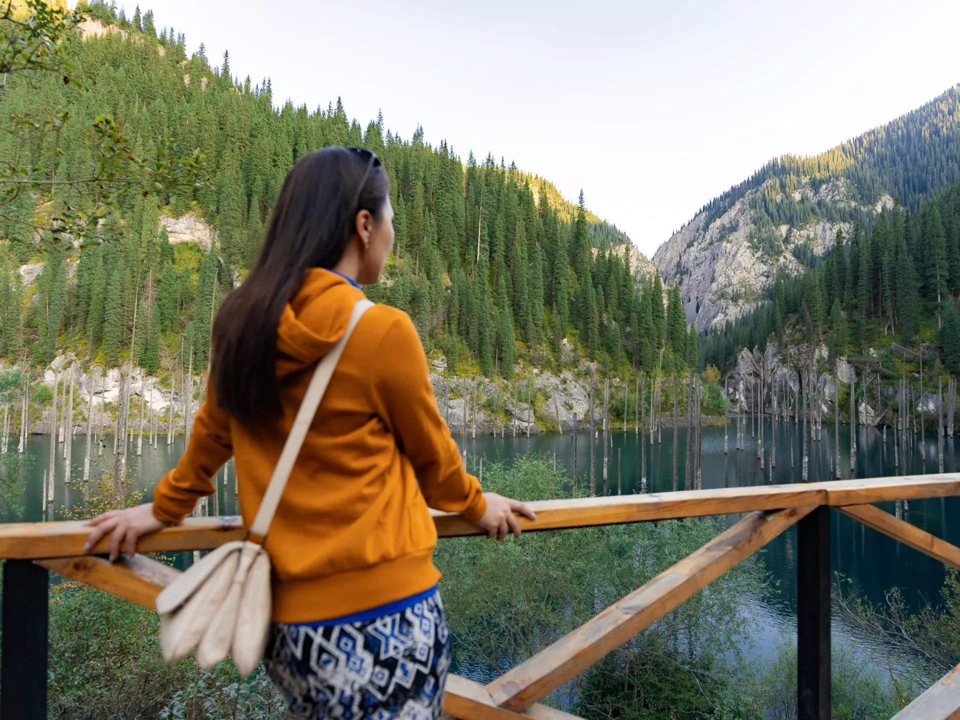 A person wearing an orange top and blue patterned skirt looks at a green lake in a valley