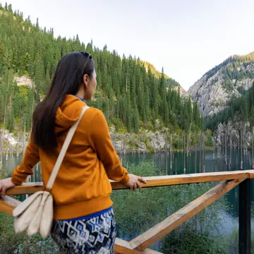 A person wearing an orange top and blue patterned skirt looks at a green lake in a valley