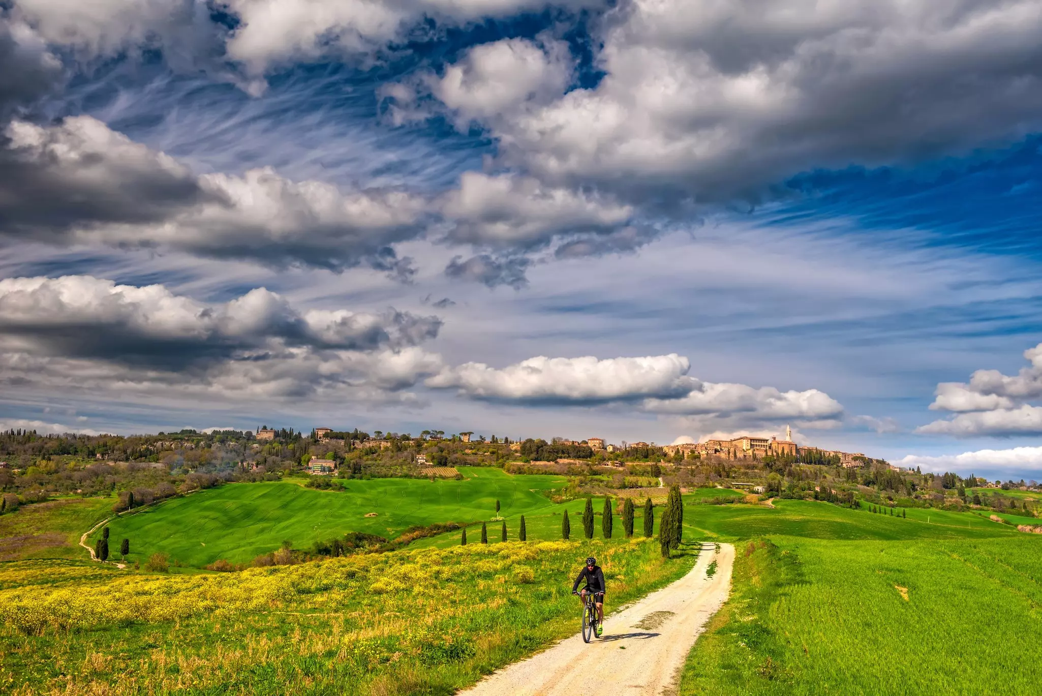 A cyclist pedals along a winding path through countryside with a small town on a distant hilltop