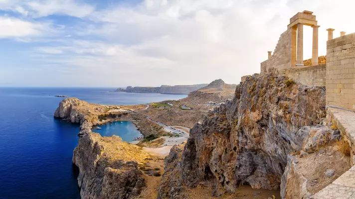 The ruins of a columned building perches on the edge of a rocky hill with the blue sea in the background. 