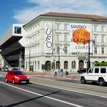 Cream neoclassical building hung with exhibition banners beside a modern cantilevered wing, with cars on the road in front.