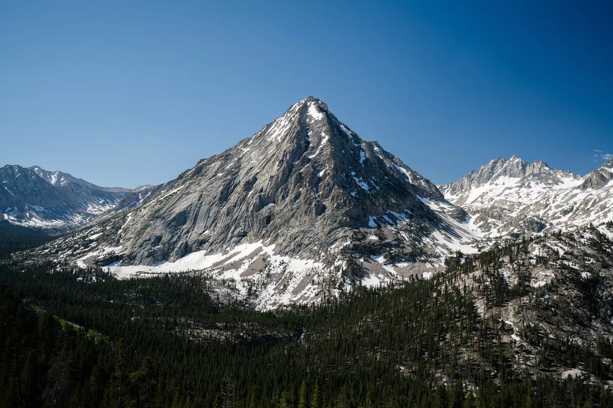 A pointed mountain peak with snow near its base where the mountain meets the tree line.