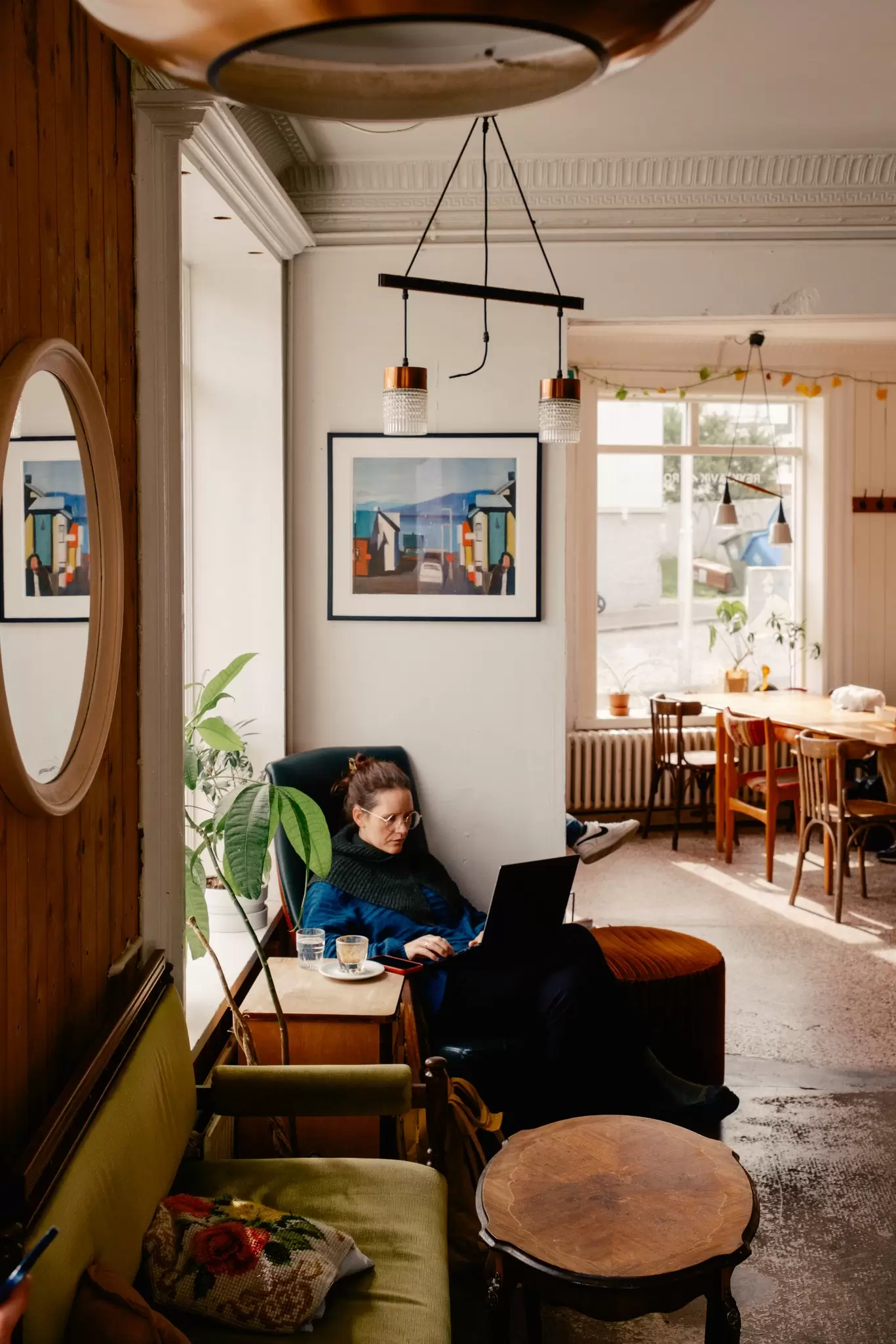 A woman sits with her laptop open on her lap in a big cozy armchair in a cafe.
