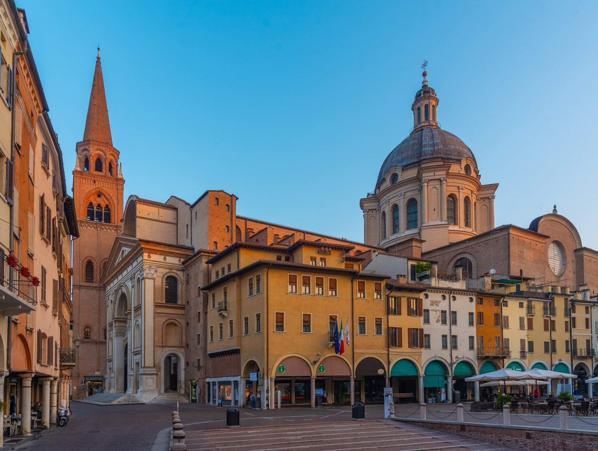 A brick bell tower and large dome rise behind ochre buildings, shops and cafe tables on a quiet square in Mantua, Italy.