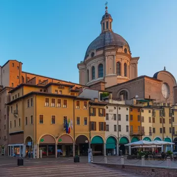 A brick bell tower and large dome rise behind ochre buildings, shops and cafe tables on a quiet square in Mantua, Italy.
