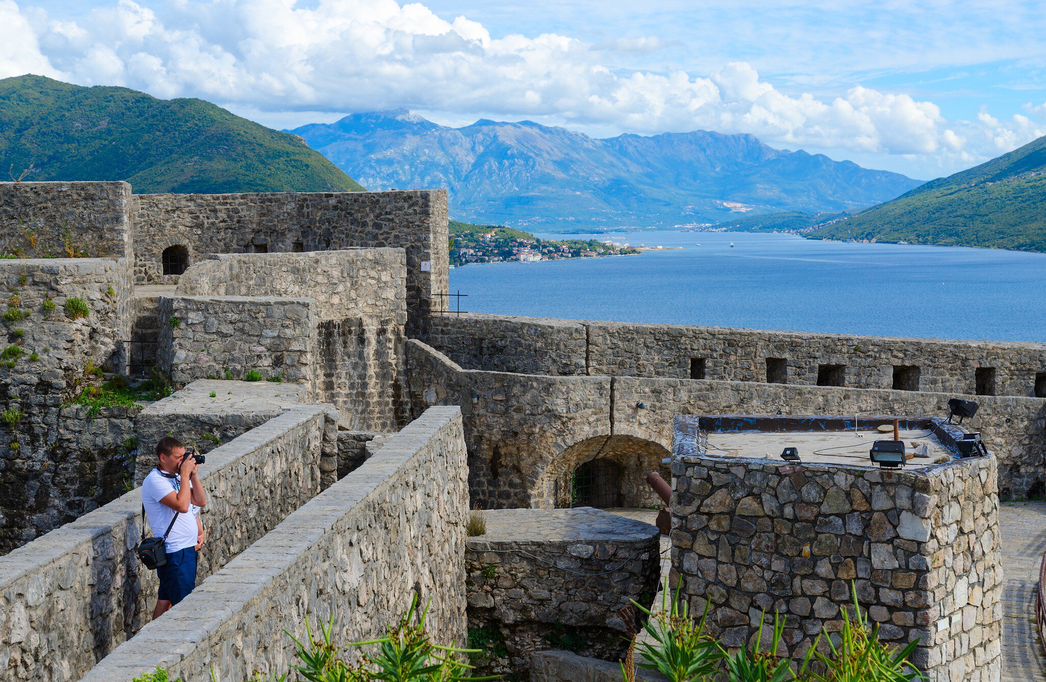 Grey stone fortress walls and an arched gateway above a blue bay, green mountains beyond, a man taking a photo.