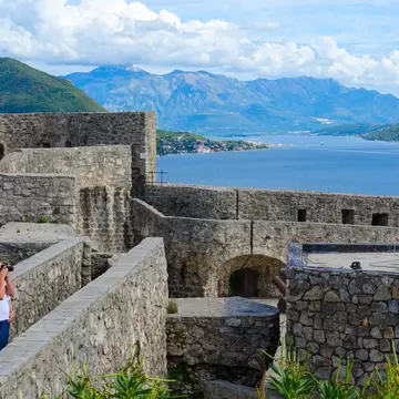 Grey stone fortress walls and an arched gateway above a blue bay, green mountains beyond, a man taking a photo.