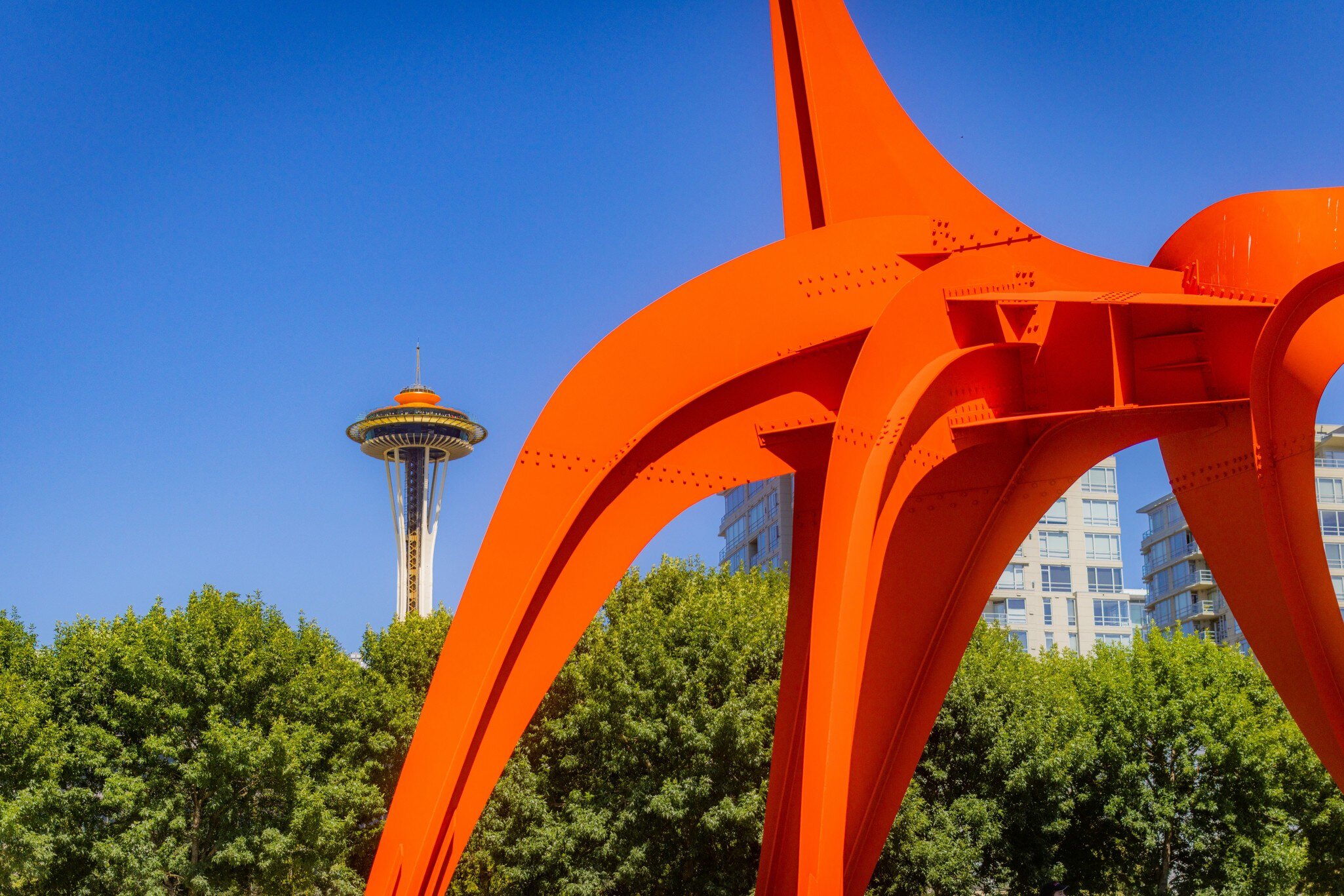 Orange steel legs of a sculpture arc across a cloudless sky, framing the distant Space Needle above green trees.