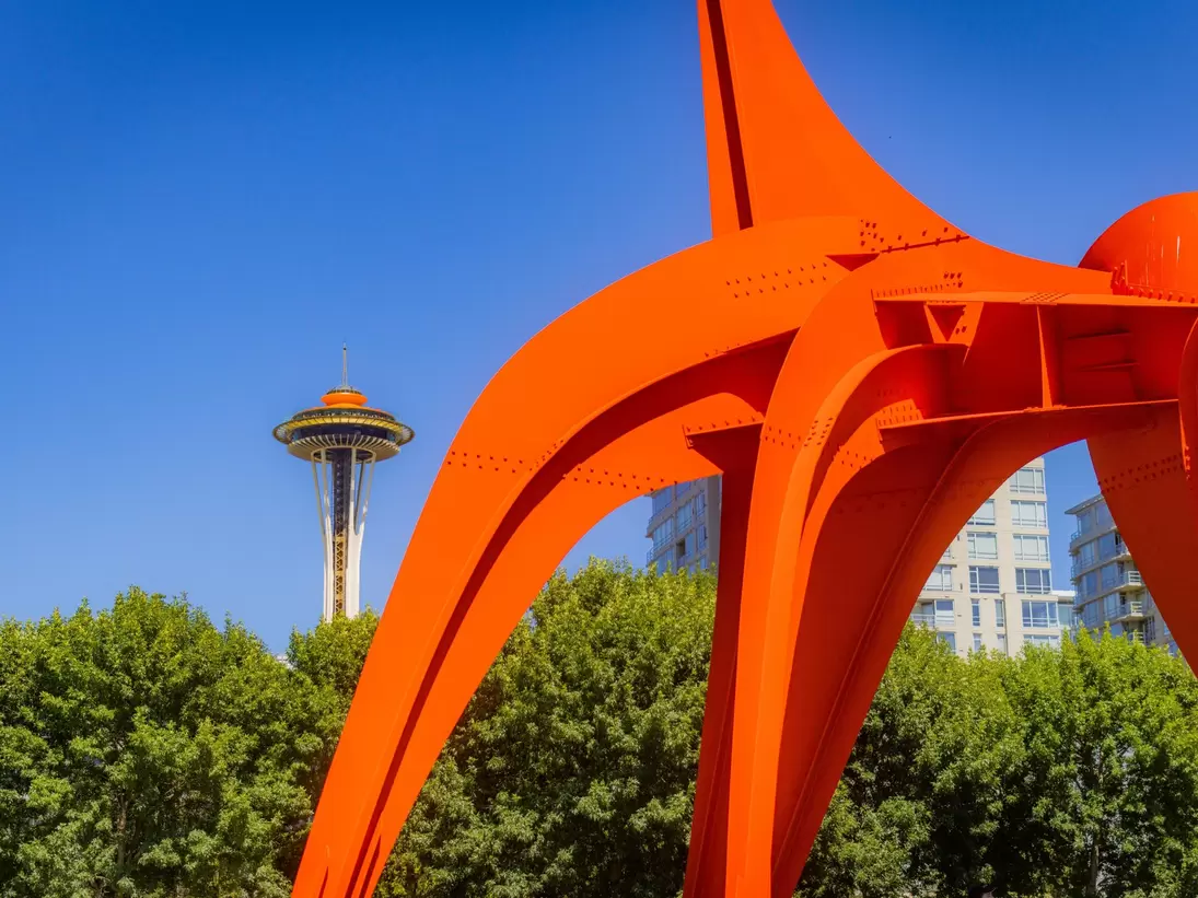 Orange steel legs of a sculpture arc across a cloudless sky, framing the distant Space Needle above green trees.