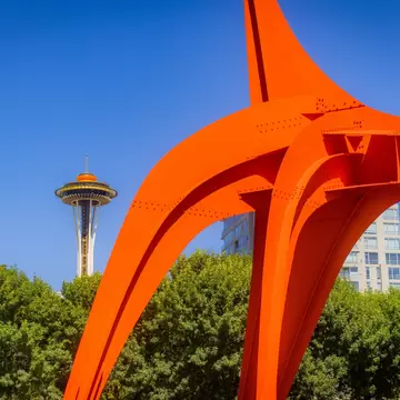 Orange steel legs of a sculpture arc across a cloudless sky, framing the distant Space Needle above green trees.