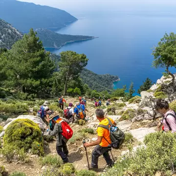 Hikers with backpacks walking down a rocky trail among pines and shrubs, blue sea and headlands below, near Fethiye