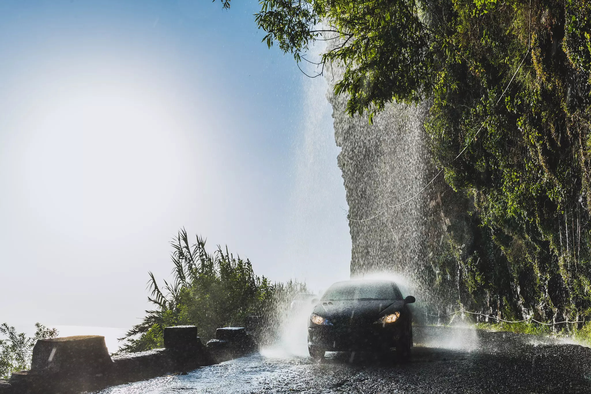 A car is seen getting drenched by a small waterfall that falls onto a road along the coast of an island.