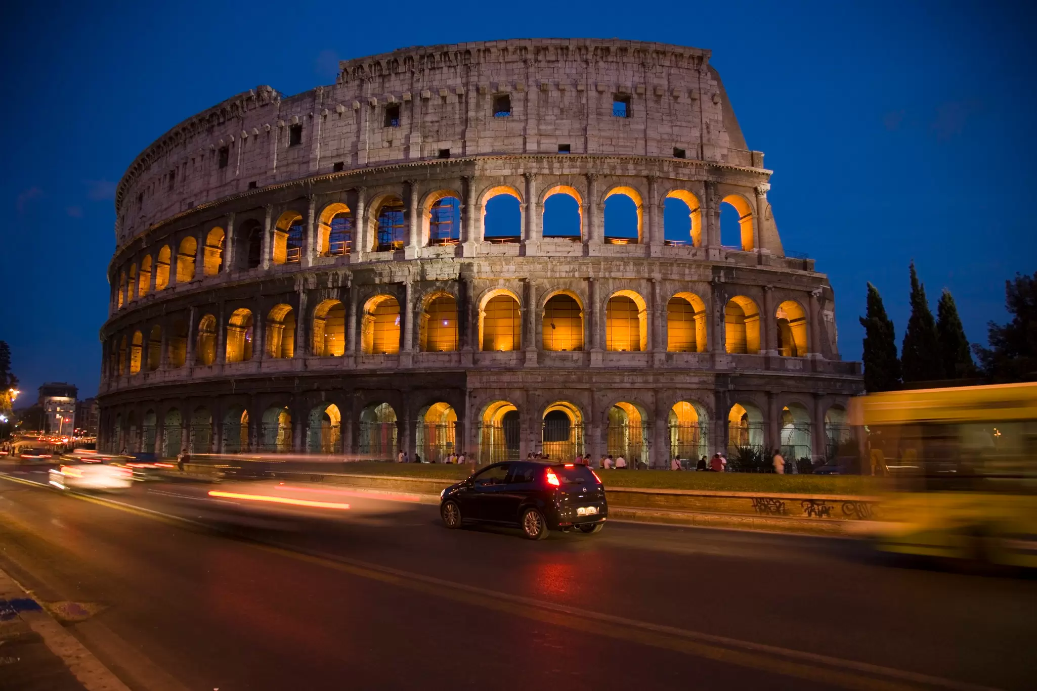 The Colosseum in Rome glowing golden at dusk against a deep blue sky, with car light trails on the road below