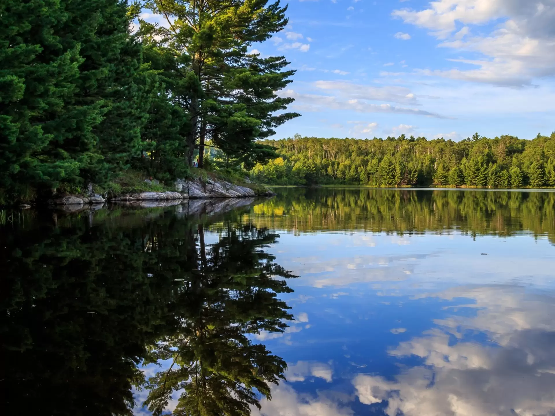 Clouds and blue sky are reflected in a calm lake with a forested shore.