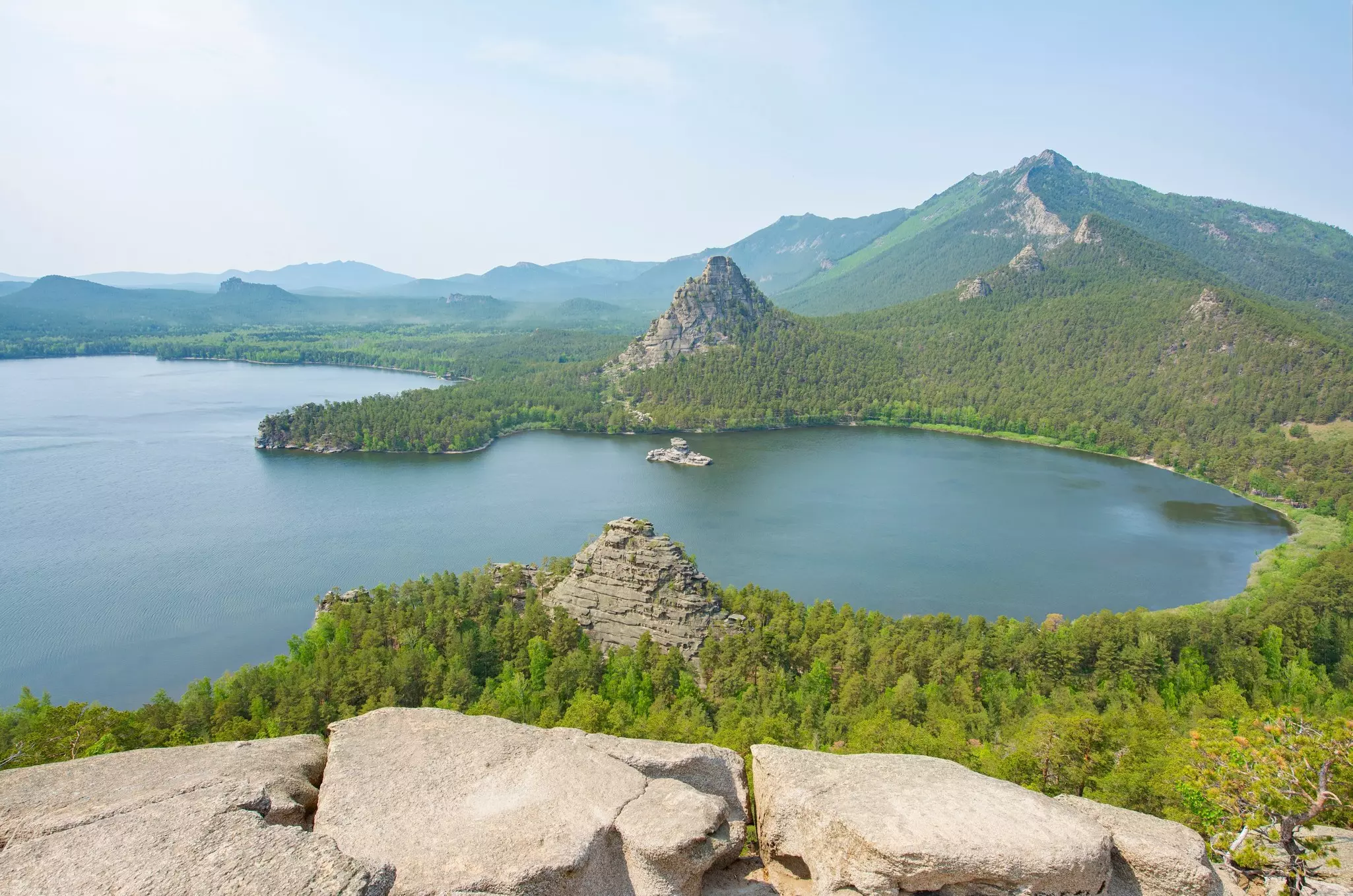 An aerial view of the curved cove of a lake, with greenery on the shoreline and several rocky outcrops.