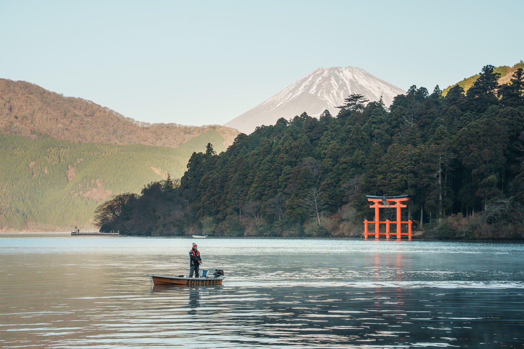 Angler in a small boat on Lake Ashi in Hakone, a red torii gate on the shore and snowy Mt Fuji behind