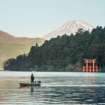 Angler in a small boat on Lake Ashi in Hakone, a red torii gate on the shore and snowy Mt Fuji behind