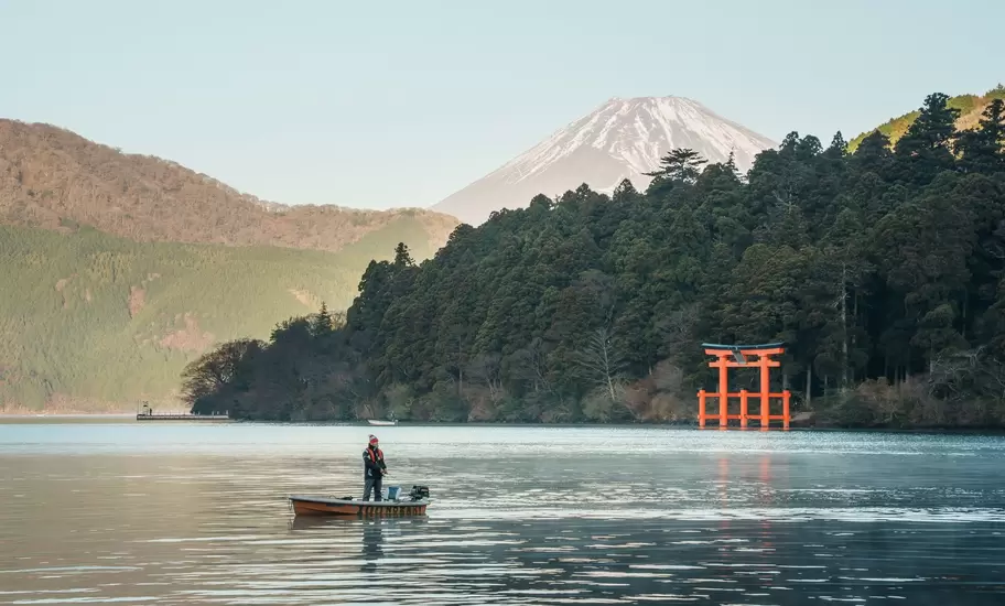 Angler in a small boat on Lake Ashi in Hakone, a red torii gate on the shore and snowy Mt Fuji behind