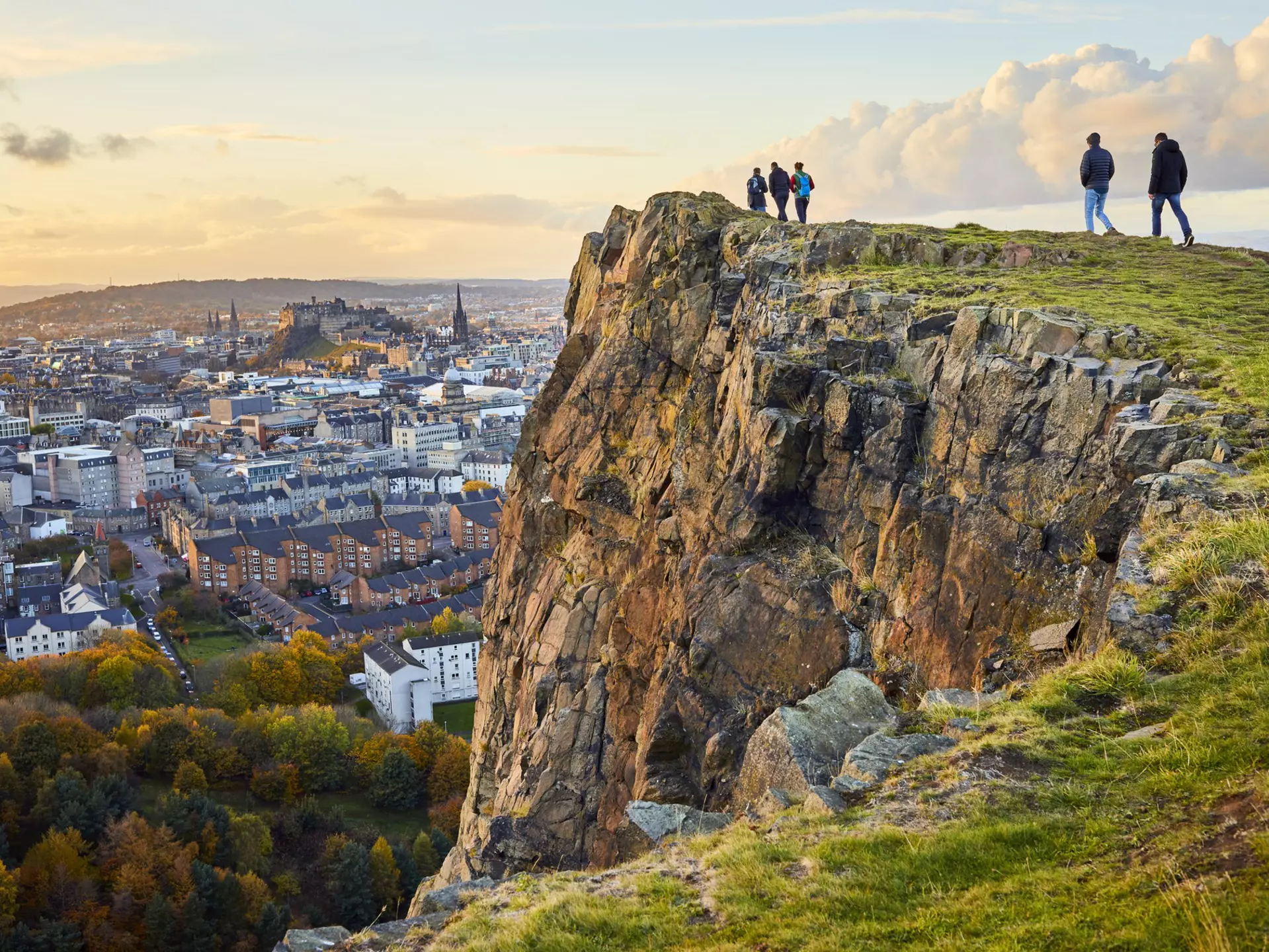 Group of people walking along cliff edge looking at city views. Edinburgh Castle in the distance