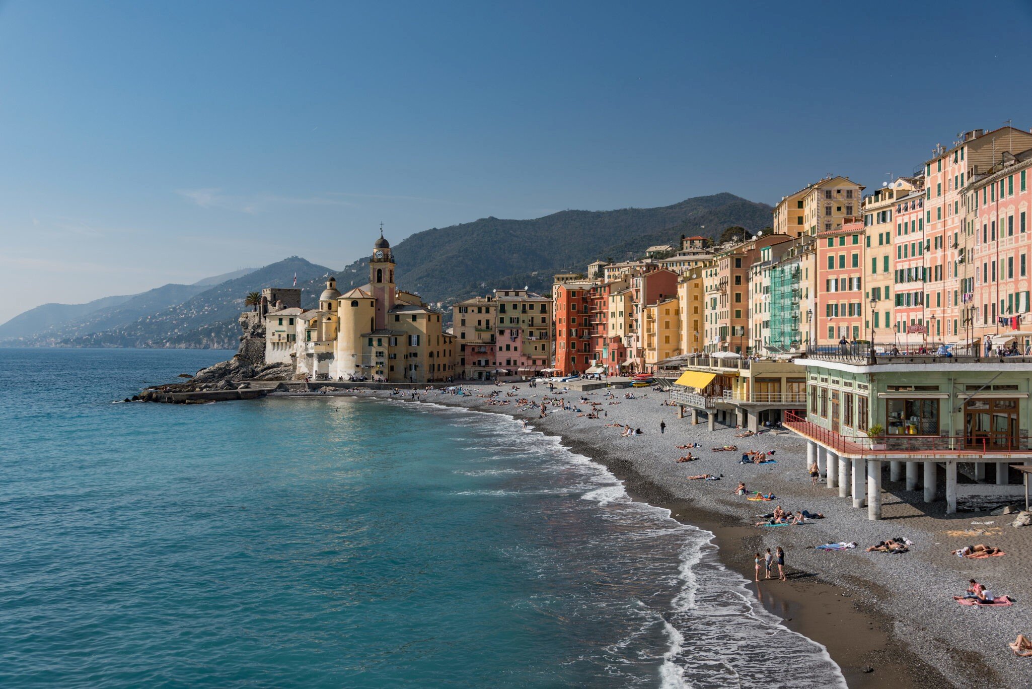 A curving pebble beach with sunbathers below tall pastel-coloured houses and a church, with green hills behind.