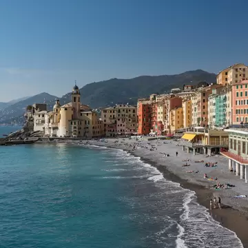 A curving pebble beach with sunbathers below tall pastel-coloured houses and a church, with green hills behind.