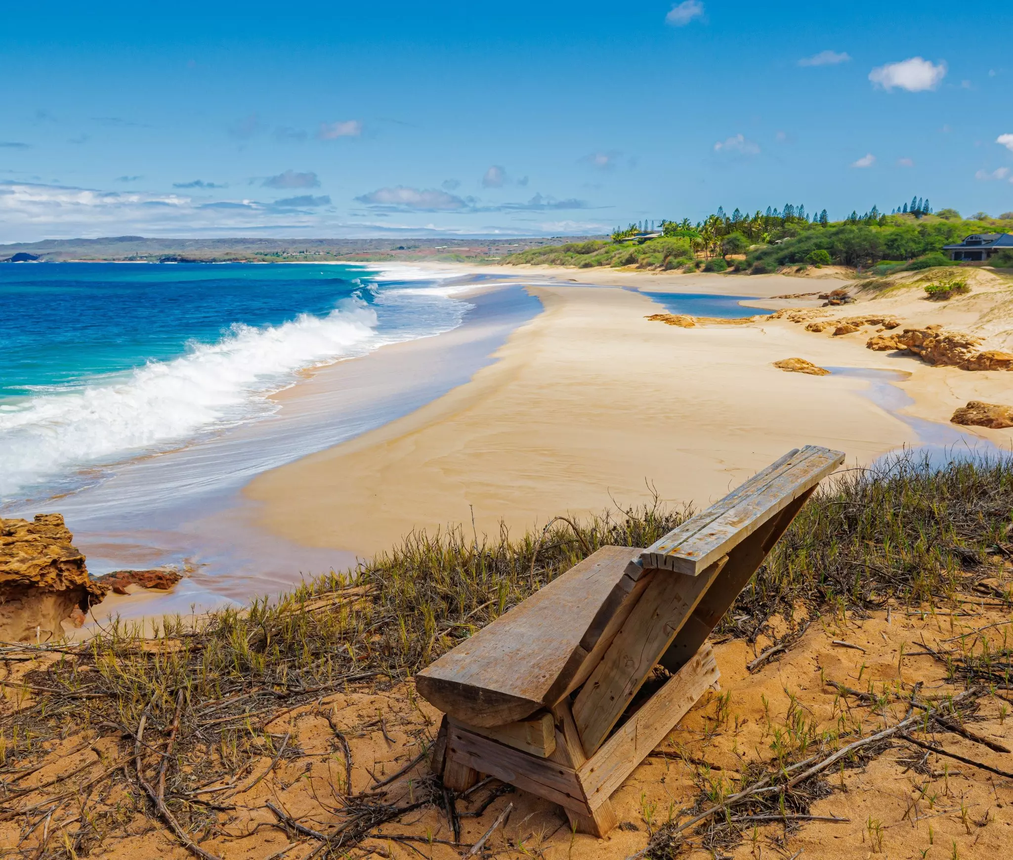 A wooden lounge chair is pictured on a dune overlooking a wide, empty beach