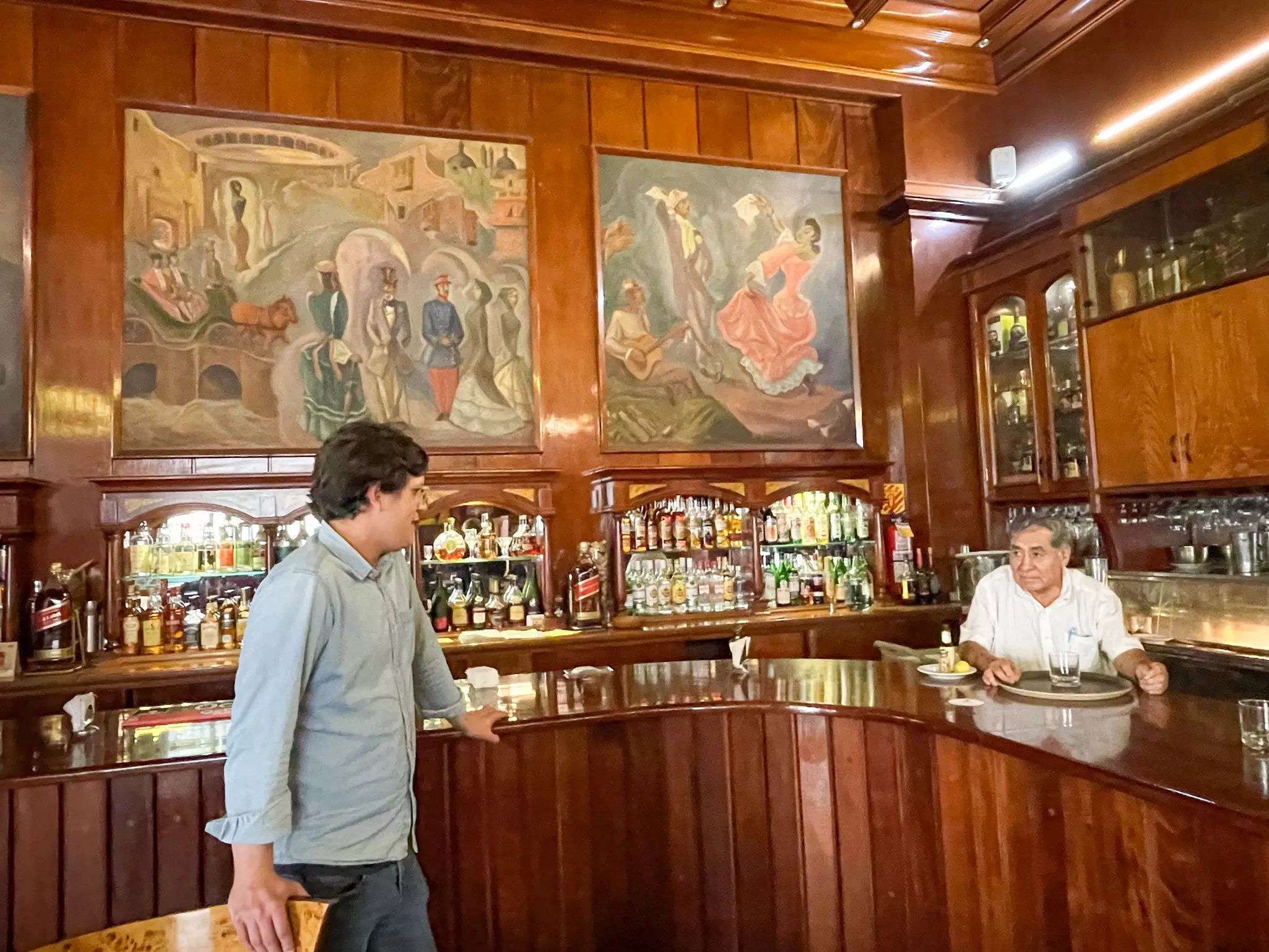 A man leans against a wood bar in Peru; the bartender is at the other end. There are bottles displayed behind the bar and murals above the shelves.