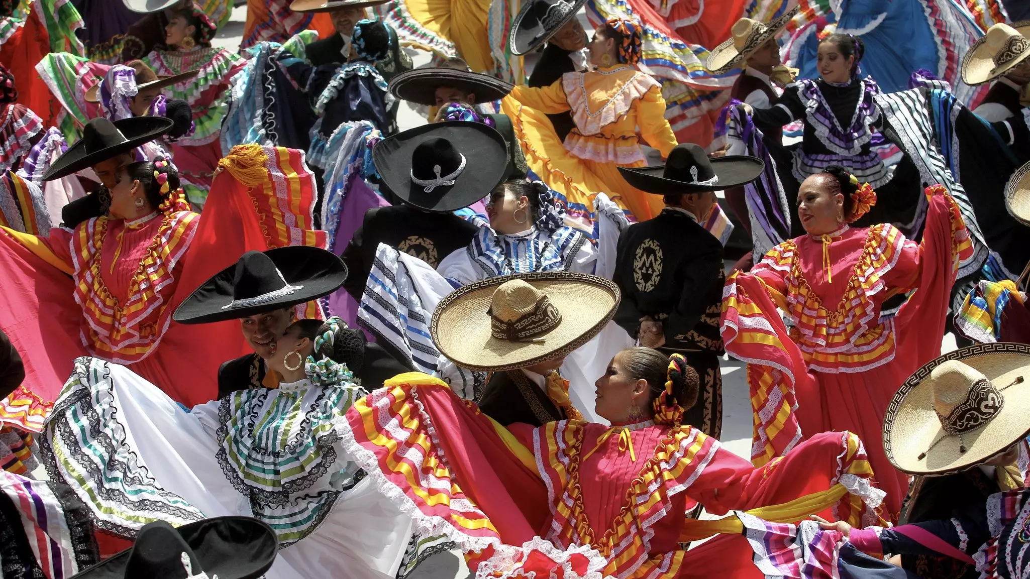 A tightknit group of dancers, with women in colorful dresses and men in dark suits with large black hats, dance together 
