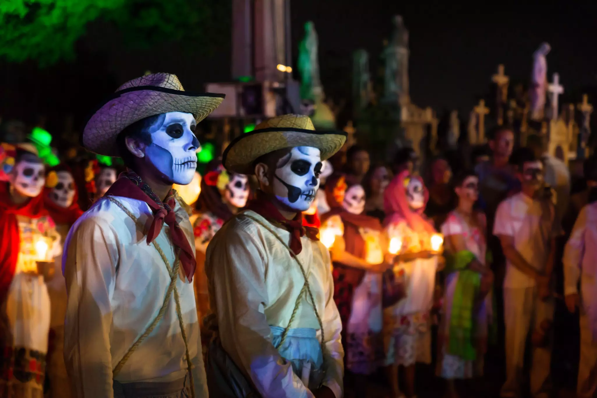 Two costumed cowboys with skull make-up in front of a line of costumed people with candles and graveyard at the event for dia de muertos