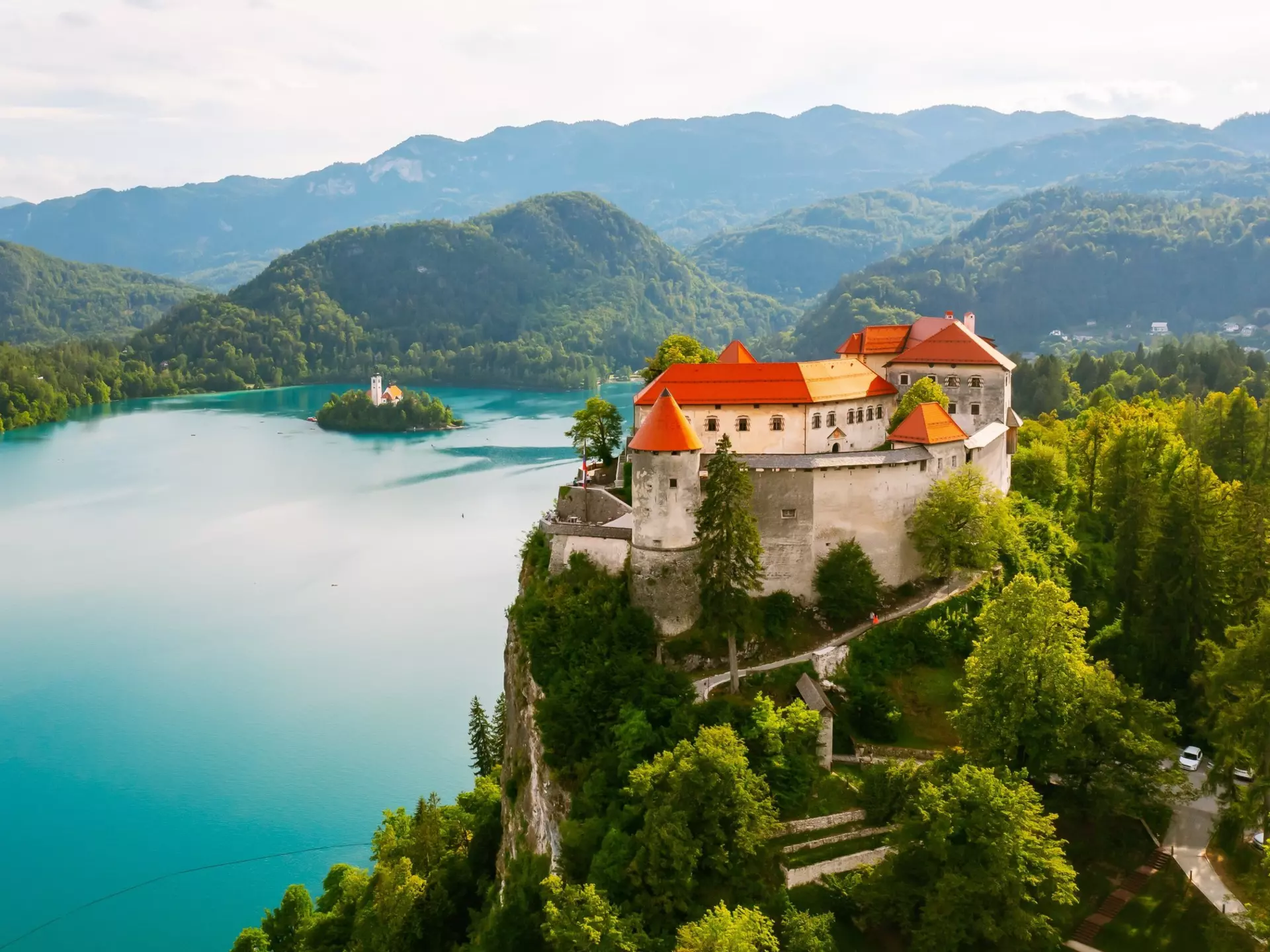 A castle with a red roof on a green hill above a blue lake that has an island in the middle with a church.