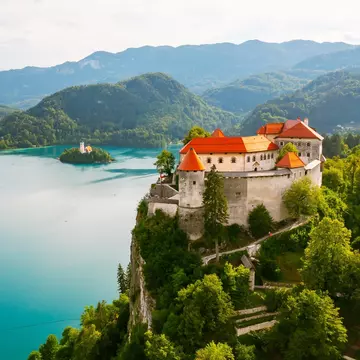 A castle with a red roof on a green hill above a blue lake that has an island in the middle with a church.