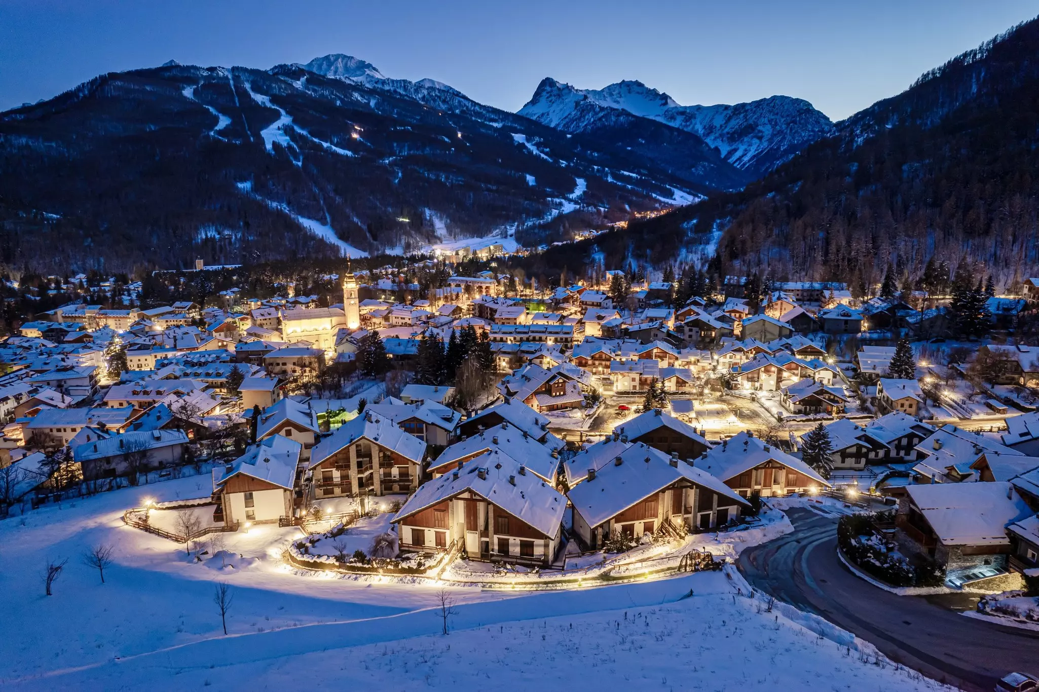A panoramic view of a village with chalets at night. Snow covered the ground and the roofs. Ski trails on mountains are visible in the distance.
